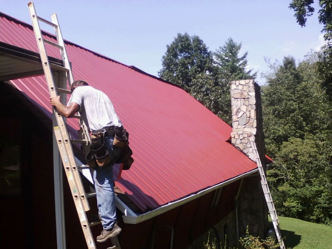 Roofer on a ladder beside a red metal roof and stone chimney — Soffit & Fascia Repair by Roofing Brothers in Smartsville